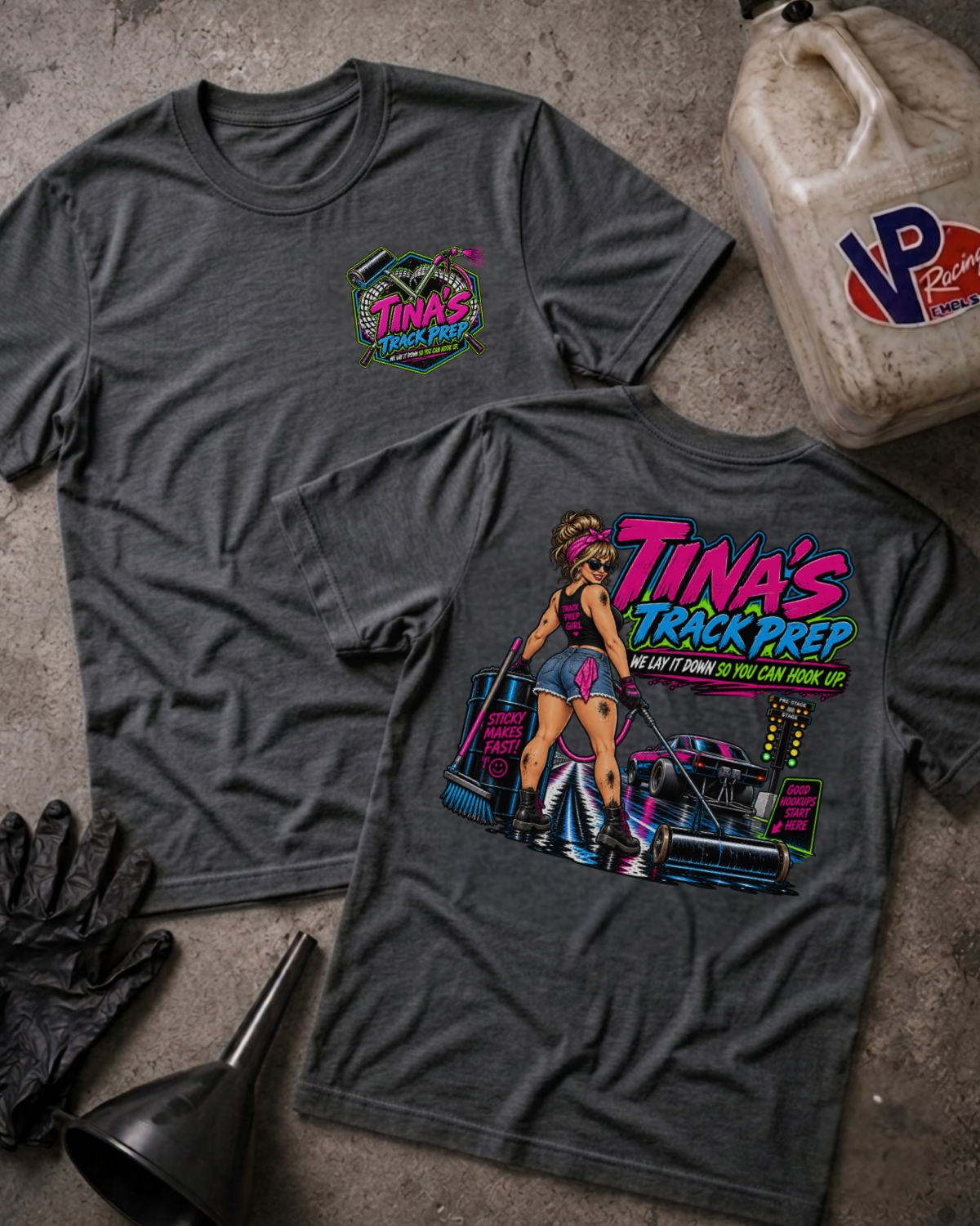 Two dark gray graphic t-shirts laid flat on a concrete surface, with a black funnel, black gloves, and a plastic jug nearby. The front shirt shows a small colorful chest graphic reading 'Tina's Track Prep', and the back shirt shows a large multicolor graphic with the same text and an illustrated woman beside a race car.