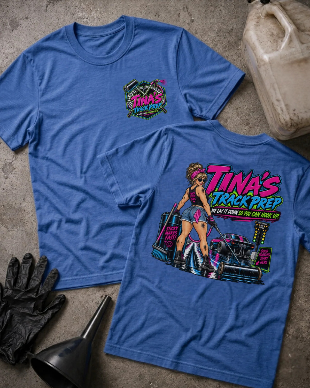 Two heather true royal graphic T-shirts laid flat on a textured concrete surface, with a gas can, black gloves, and a funnel nearby. The front shirt shows a small multicolor chest graphic reading 'Tina's Track Prep,' and the back shirt shows a large colorful graphic with the same text plus an illustrated woman and a race car.