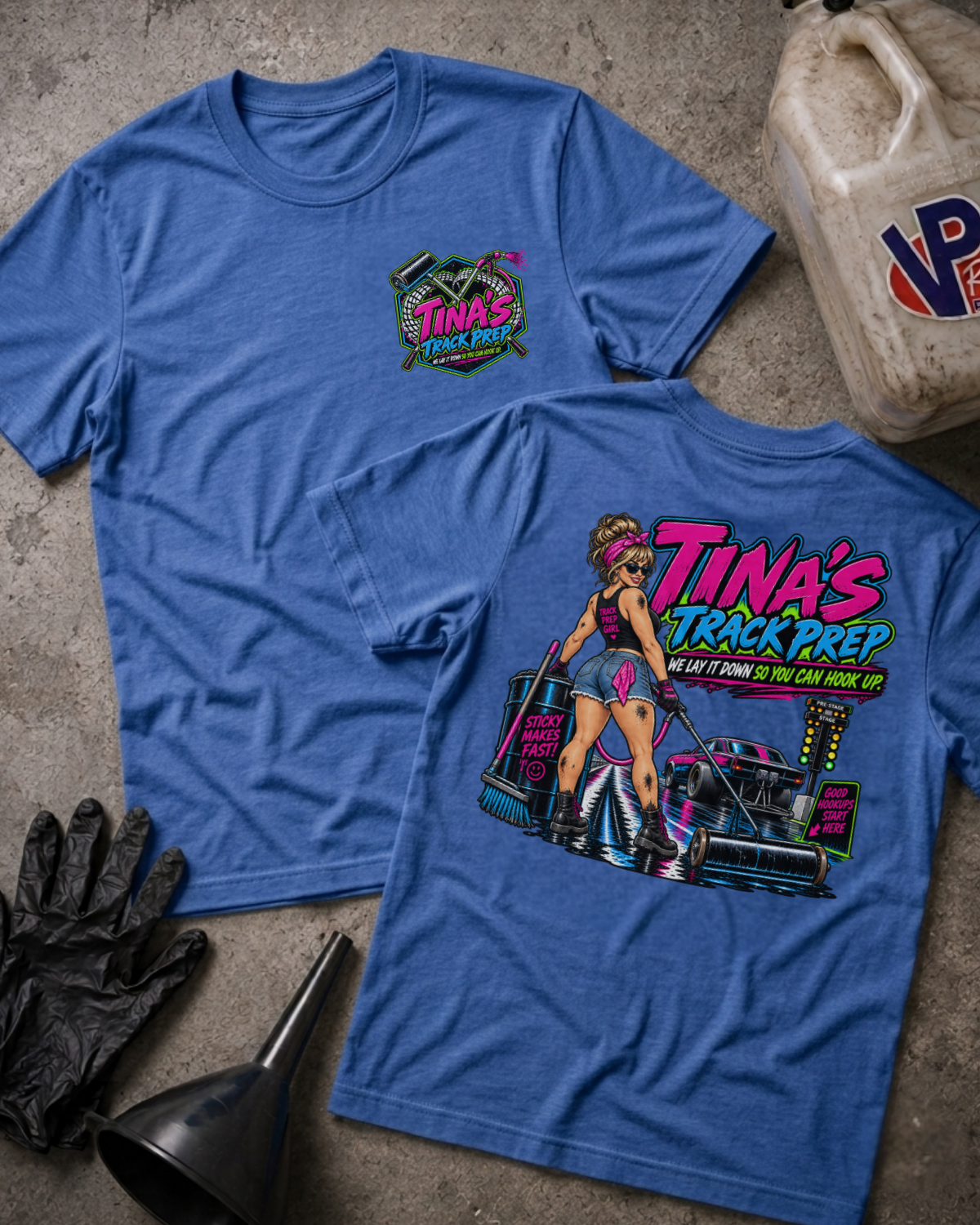 Two heather blue graphic T-shirts laid flat on a concrete surface, with a black glove, funnel, and a worn plastic jug nearby. The front shirt shows a small multicolor chest graphic reading Tina's Track Prep, and the back shirt shows a large colorful graphic of a pin-up woman with a race car and the text 'TINA'S TRACK PREP' plus 'WE LAY IT DOWN SO YOU CAN HOOK UP'.