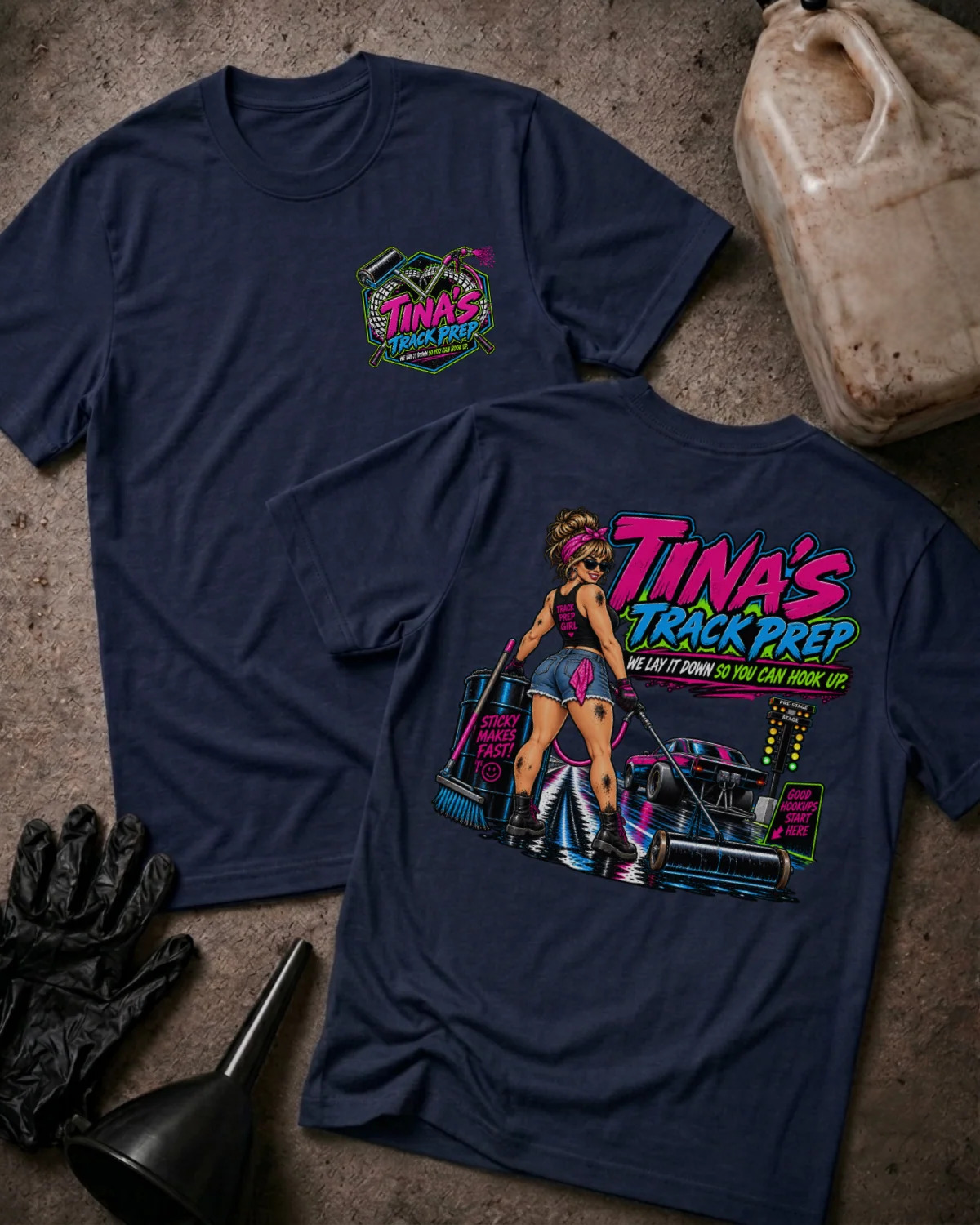 Two navy graphic T-shirts laid flat on a textured concrete surface, with a gas can, black gloves, and a funnel nearby. The front shirt shows a small multicolor chest graphic reading 'Tina's Track Prep,' and the back shirt shows a large colorful graphic with the same text plus an illustrated woman and a race car.