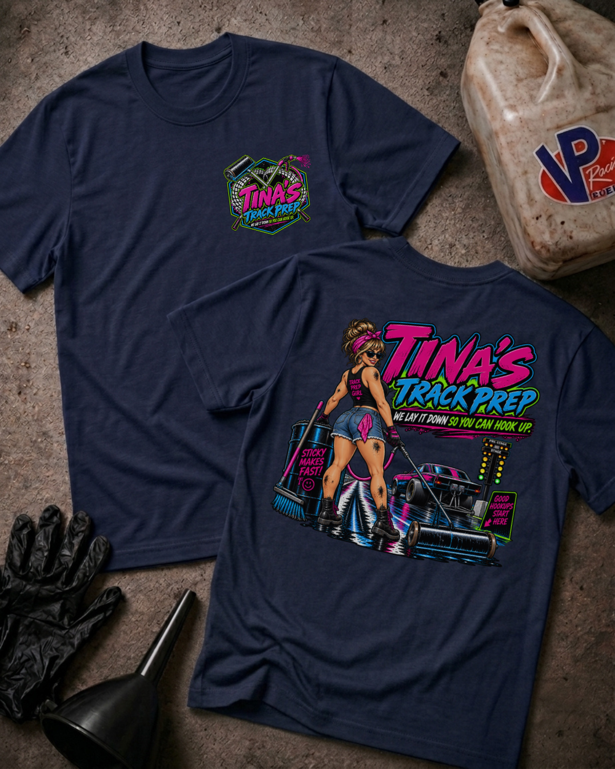 Two navy blue t-shirts displayed on a textured concrete surface, styled with a black oil pan, black gloves, and a plastic jug. The front shirt features a small colorful 'Tina's Track Prep' graphic on the left chest, while the back shirt displays a large, detailed graphic of a woman in a pink racing outfit next to a race car with the text 'TINA'S TRACK PREP'.