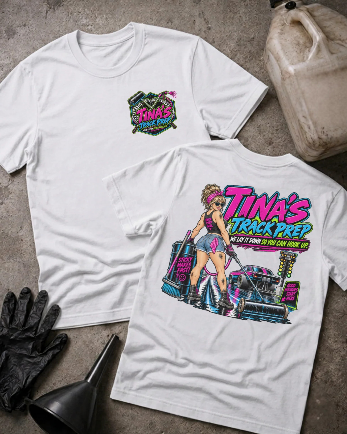 Two white graphic T-shirts laid flat on a textured concrete surface, with a gas can, black gloves, and a funnel nearby. The front shirt shows a small multicolor chest graphic reading 'Tina's Track Prep,' and the back shirt shows a large colorful graphic with the same text plus an illustrated woman and a race car.