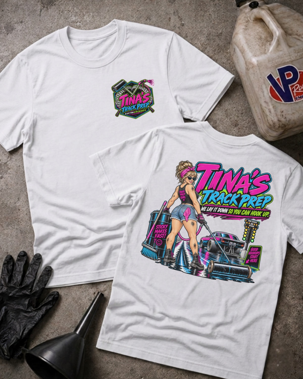 Two white graphic T-shirts laid flat on a concrete surface, showing the front and back designs. The front has a small colorful chest graphic reading 'Tina's Track Prep,' and the back has a large illustrated graphic with the same text plus a race car and a woman in athletic wear.