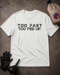 A white short-sleeve t-shirt with the text 'TOO FAST TOO FED UP' printed in black across the chest, laid flat on a dark textured surface with black gloves, a funnel, and a VP Fast Fuel tank jug nearby.