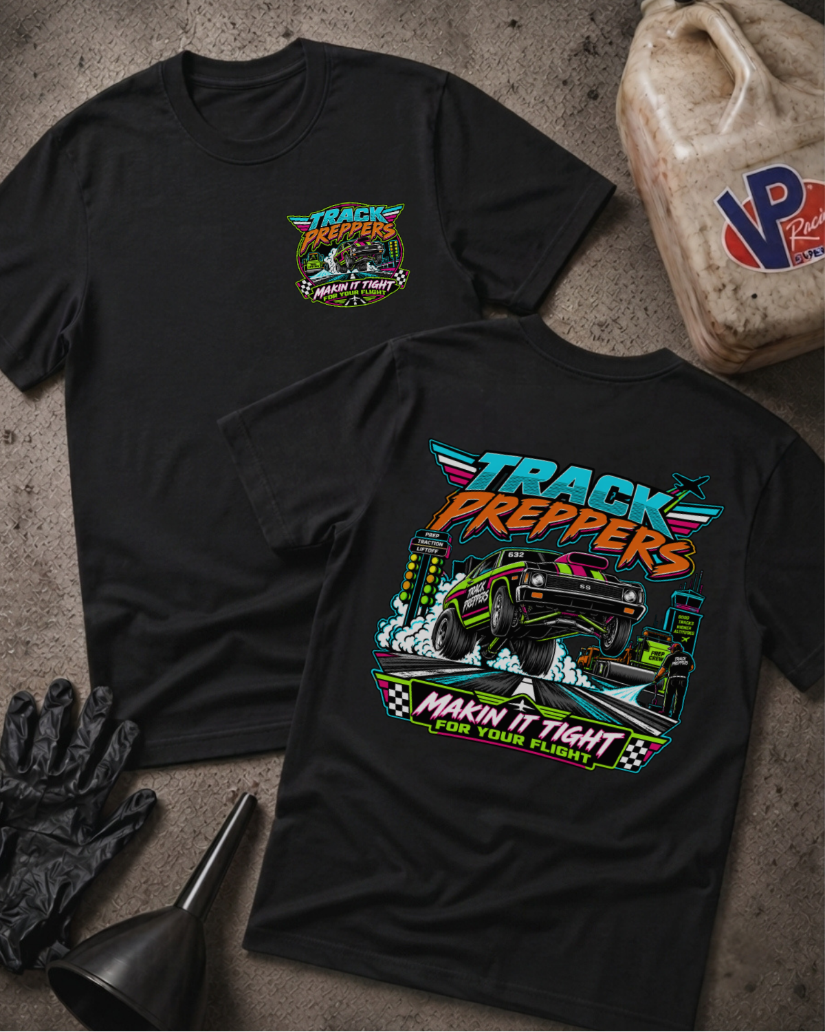 Two black graphic T-shirts displayed on a textured concrete surface, with a black funnel, black gloves, and a worn plastic jerrycan nearby. The front shirt shows a small multicolor chest graphic reading 'Track Preppers' with a monster truck illustration.
