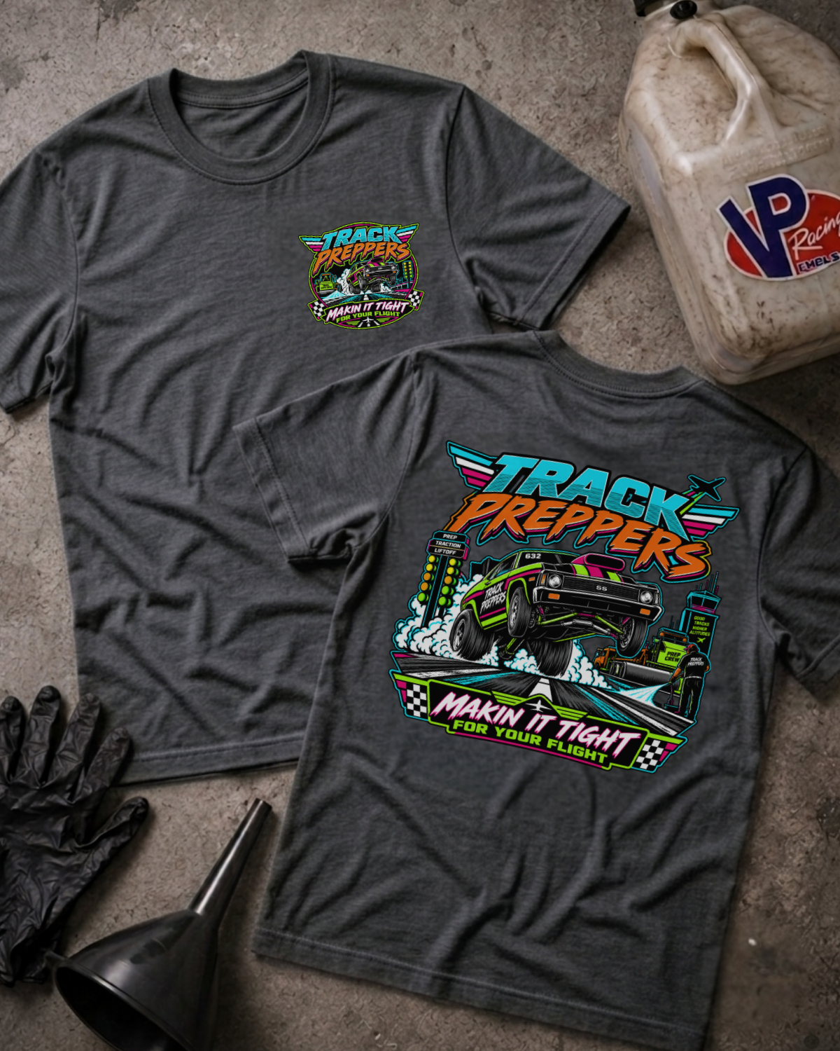 Two dark heather gray graphic T-shirts laid flat on a concrete surface, showing the front and back designs. The front has a small chest graphic reading 'TRACK PREPPERS' with a race car illustration.