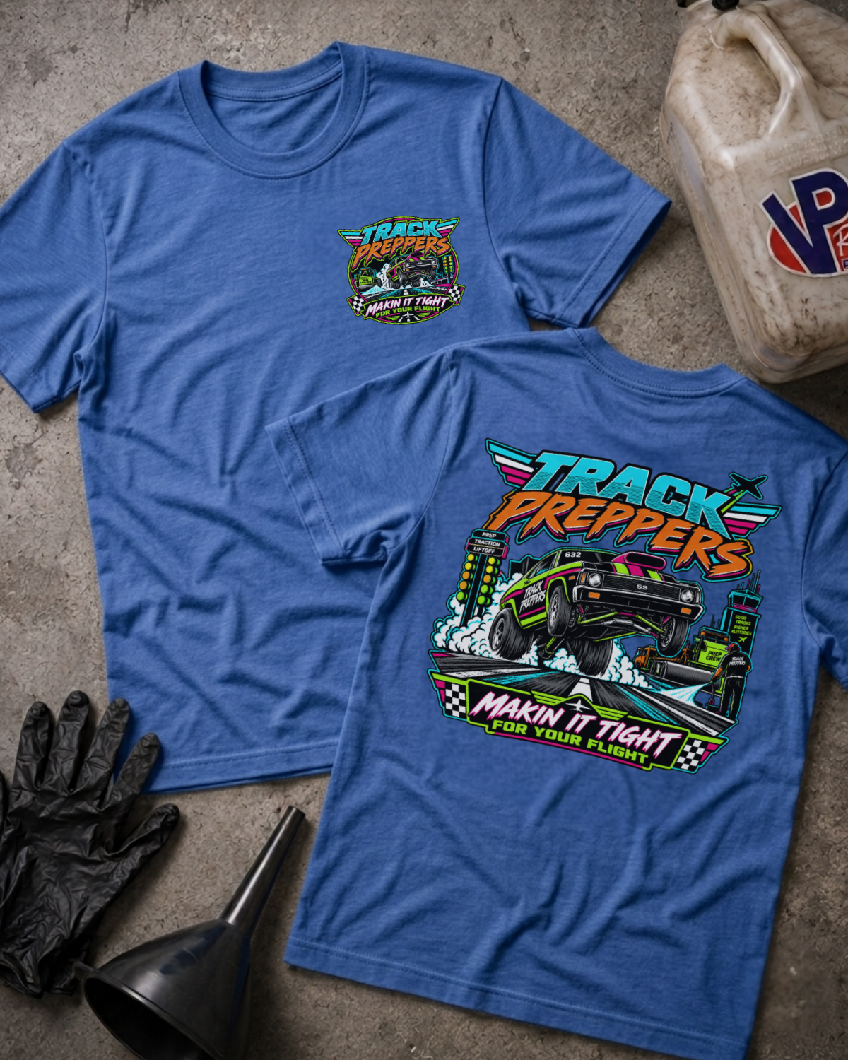Two blue graphic T-shirts laid flat on a concrete-like surface, showing the front and back designs. The shirts feature a colorful race car illustration with the text 'TRACK PREPPERS' and 'MAKIN' IT TIGHT FOR YOUR FLIGHT.