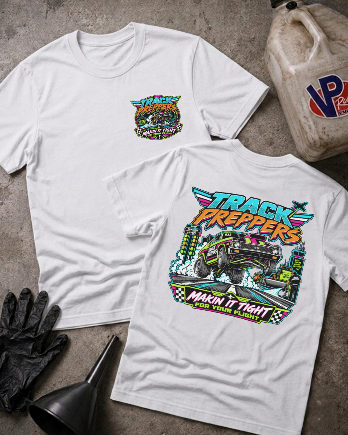 Two white graphic T-shirts laid flat on a gray concrete surface, with a black glove, funnel, and a worn fuel jug nearby. The front shirt shows a small multicolor chest graphic reading Track Preppers, and the back shirt shows a large multicolor track-themed graphic with a race car and the words Track Preppers and Makin It Tight For Your Flight.