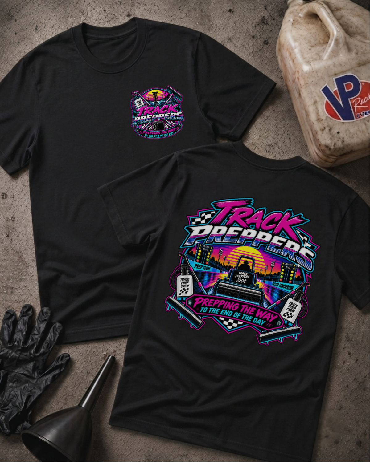 Two black graphic T-shirts laid flat on a textured gray surface, with a gas can, black gloves, and a funnel as styled props. The front shirt shows a small multicolor chest graphic reading 'Track Preppers,' and the back shirt shows a large matching multicolor graphic with neon-style lettering and racing imagery.