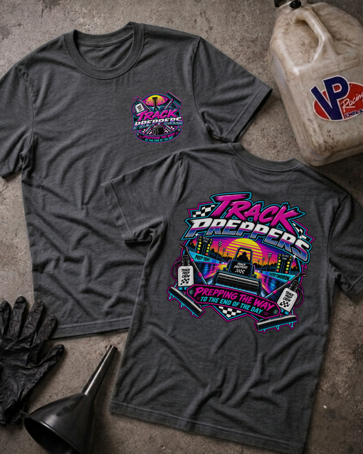 Two dark grey heather graphic t-shirts displayed flat on a textured concrete surface, surrounded by motorcycle-themed props including a gas can, gloves, and a funnel. The front shirt features a small circular logo on the left chest, while the back shirt displays a large, colorful retro-style graphic with the text 'Track Preppers' and 'Prepping The Way To The End Of The Day'.