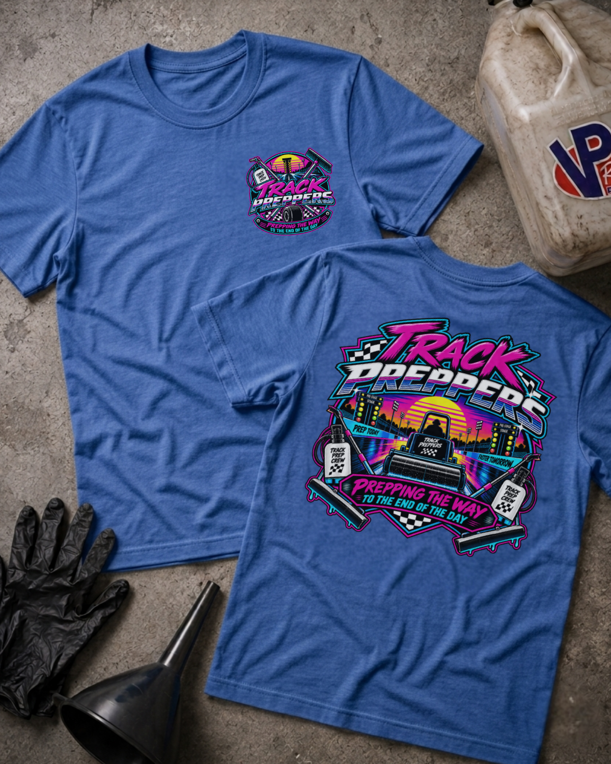 Two heather true royal graphic t-shirts displayed flat on a textured concrete surface, surrounded by motorcycle-themed props including a gas can, gloves, and a funnel. The front shirt features a small circular logo on the left chest, while the back shirt displays a large, colorful retro-style graphic with the text 'Track Preppers' and 'Prepping The Way To The End Of The Day'.