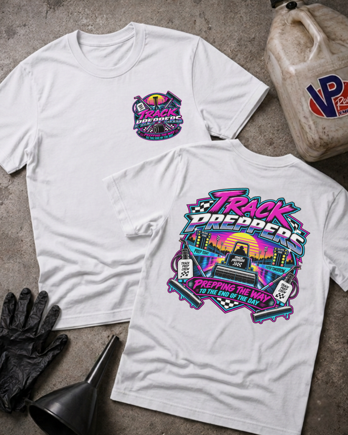 Two white graphic T-shirts displayed on a concrete floor with racing-themed props, including a black glove, funnel, and fuel jug. The front shirt shows a small chest graphic, and the back shirt shows a large colorful 'Track Preppers' graphic.