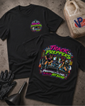 Two black graphic t-shirts displayed on a textured concrete surface with garage-themed props including a fuel jug, black gloves, and a funnel. The front shirt features a small circular 'Track Preppers' logo on the left chest, while the back shirt displays a large, colorful graphic of two women in a garage with the text 'TRACK PREPPERS' and 'PREPPING TO THE END SO YOU CAN HIT SEND'.