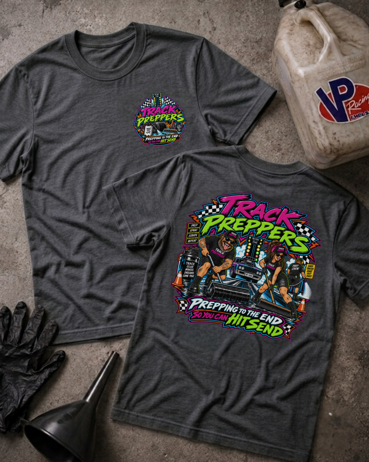 Two dark heather gray graphic t-shirts displayed flat on a concrete floor, with a black oil jug, black gloves, and a black funnel as props. The front shirt features a small colorful chest graphic, while the back shirt features a large, detailed multicolor graphic with the text 'TRACK PREPPERS' and 'PREPPING TO THE END SO I CAN HIT SEND'.