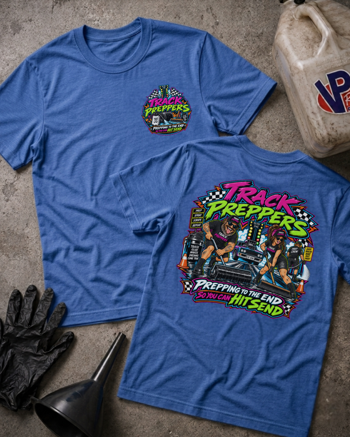 Two blue graphic T-shirts laid flat on a concrete floor with racing props, showing both front and back designs. The front has a small multicolor chest graphic reading 'Track Preppers,' and the back has a large colorful graphic with the same text and a driver at a race track.
