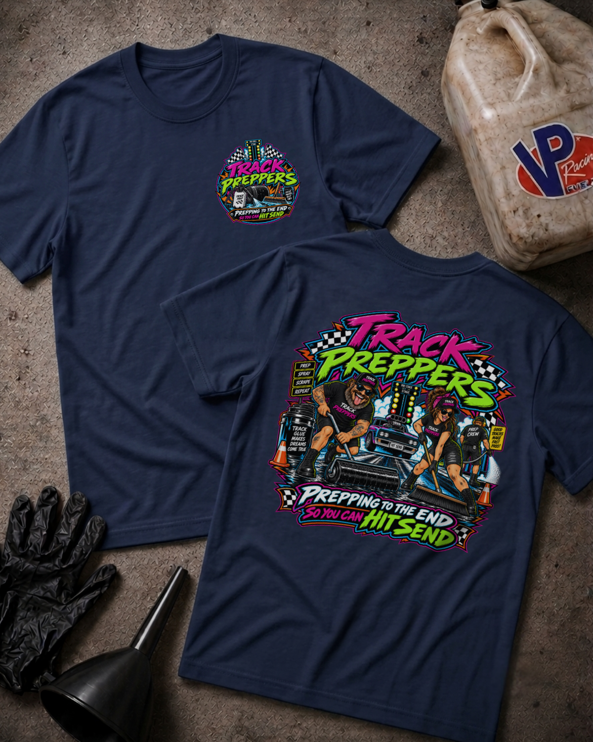 Two navy blue t-shirts displayed on a textured gray surface with garage-themed props. The front shirt shows a colorful 'Track Preppers' chest graphic, and the back shirt features a large, detailed multicolor graphic with the text 'Track Preppers' and 'Prepping to the End So You Can Hit Send'.