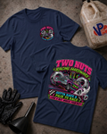 Two navy blue graphic T-shirts laid flat on a textured concrete surface, with a race gas jug, black gloves, and funnel as props. The front shirt shows a small multicolor chest graphic, and the back shirt shows a large colorful racing-themed print with a blue muscle car and the text 'TWO NUTS RACING HARDWARE' and 'GOTTA HAVE A PAIR TO RACE LOSE A NUT, WRECK YOUR BUTT'.