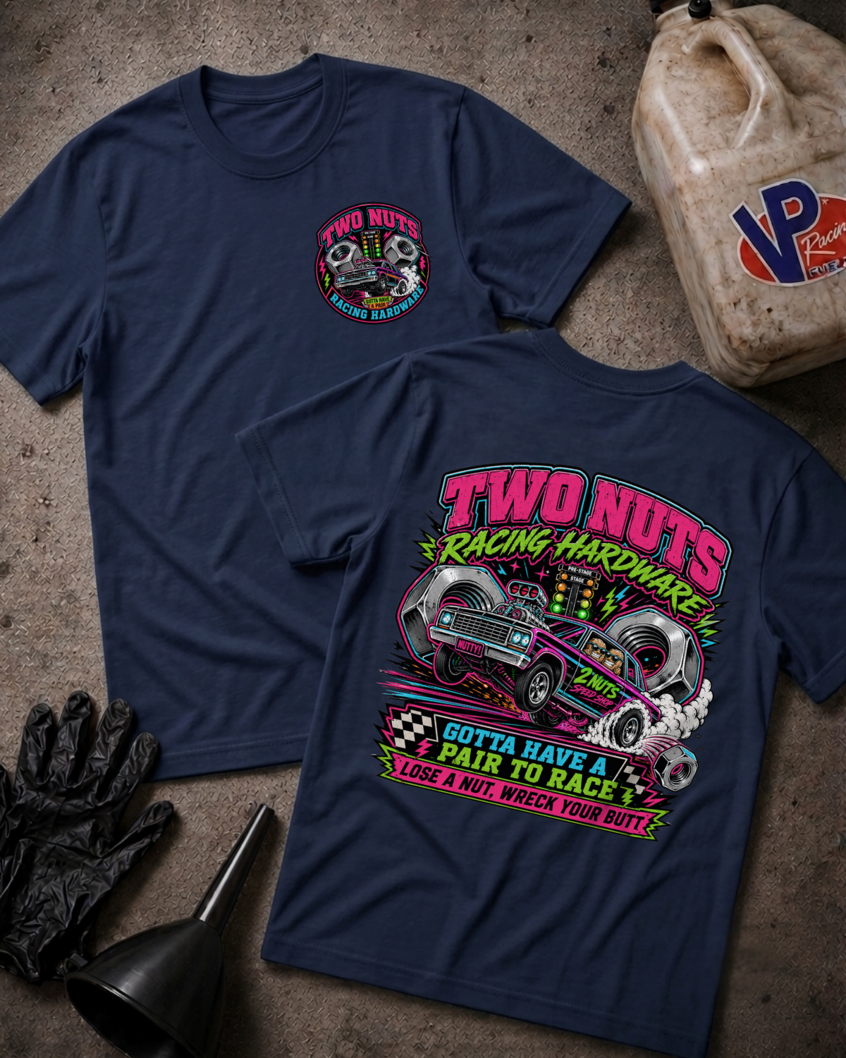 Two navy blue graphic T-shirts laid flat on a textured concrete surface, with a race gas jug, black gloves, and funnel as props. The front shirt shows a small multicolor chest graphic, and the back shirt shows a large colorful racing-themed print with a blue muscle car and the text 'TWO NUTS RACING HARDWARE' and 'GOTTA HAVE A PAIR TO RACE LOSE A NUT, WRECK YOUR BUTT'.