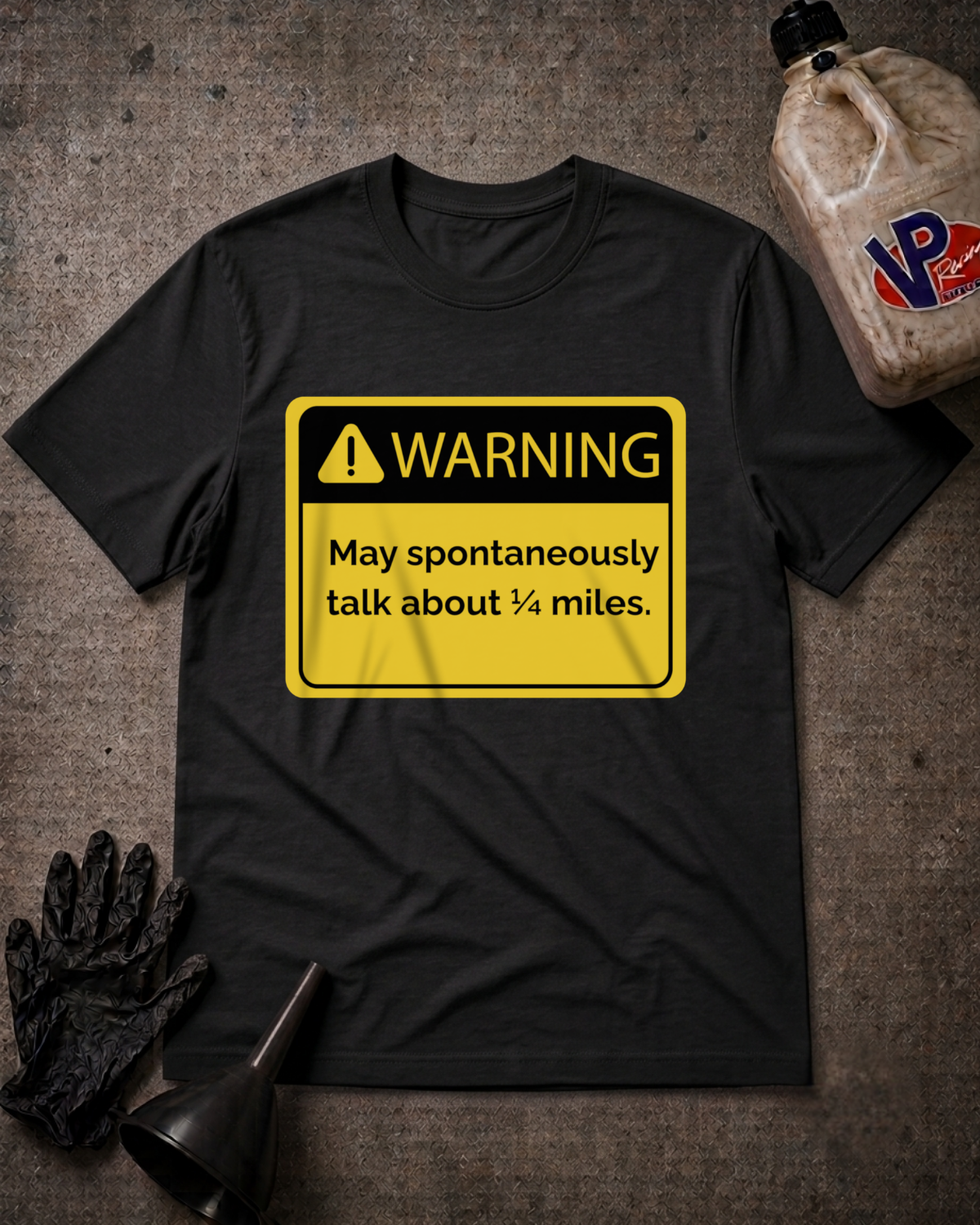 A black short-sleeve t-shirt with a yellow warning sign graphic that reads 'WARNING May spontaneously talk about 1/4 miles.' It is displayed in a flat-lay arrangement on a textured gray background, accompanied by a black work glove, a funnel, and a plastic jug in the corners.
