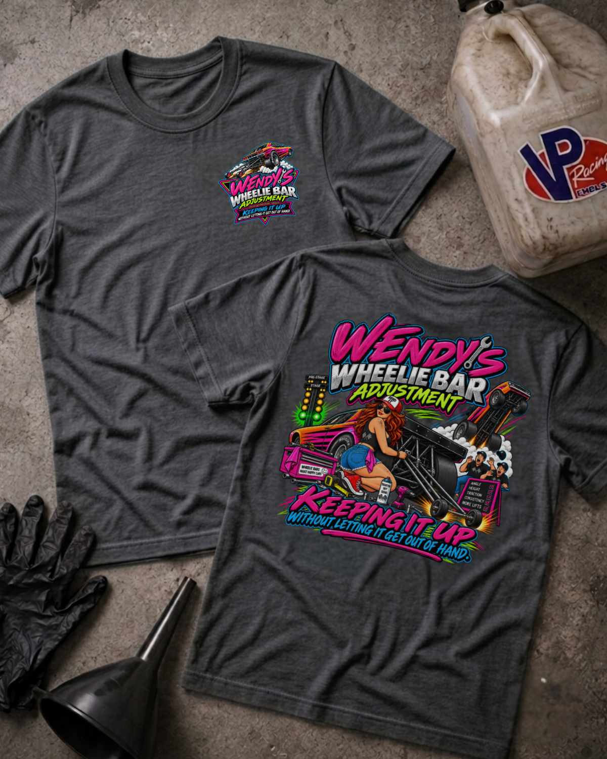 Two dark grey heather graphic t-shirts displayed on a textured concrete surface with garage-themed props including a plastic jug, black gloves, and a funnel. The front shirt shows a small pink and blue logo on the left chest, while the back shirt features a large colorful graphic of a woman in a pink car with the text 'Wendy's Wheelie Bar Adjustment Keeping It Up'.