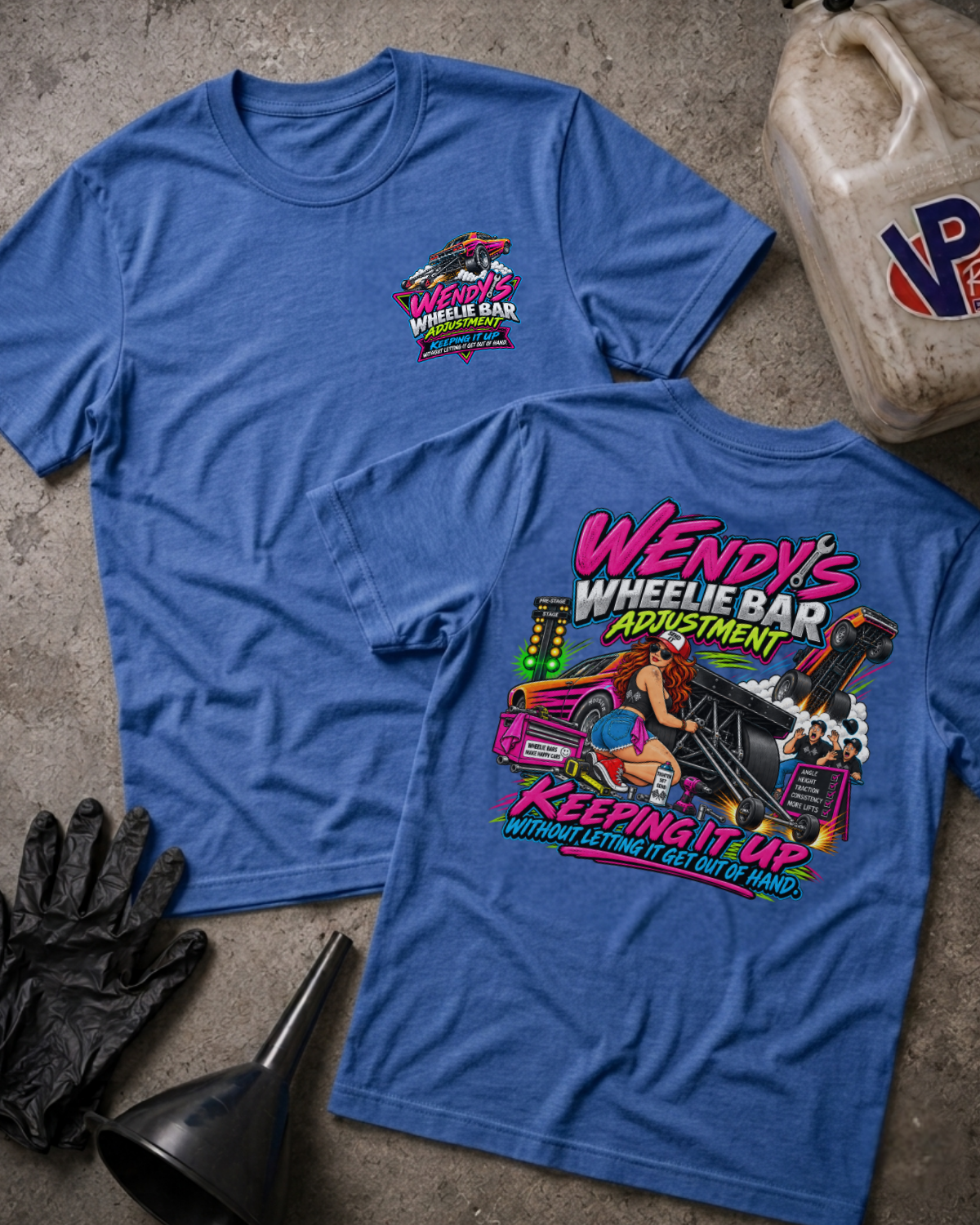 Two heather true royal graphic t-shirts displayed on a textured concrete surface with garage-themed props including a plastic jug, black gloves, and a funnel. The front shirt shows a small pink and blue logo on the left chest, while the back shirt features a large colorful graphic of a woman in a pink car with the text 'Wendy's Wheelie Bar Adjustment Keeping It Up'.
