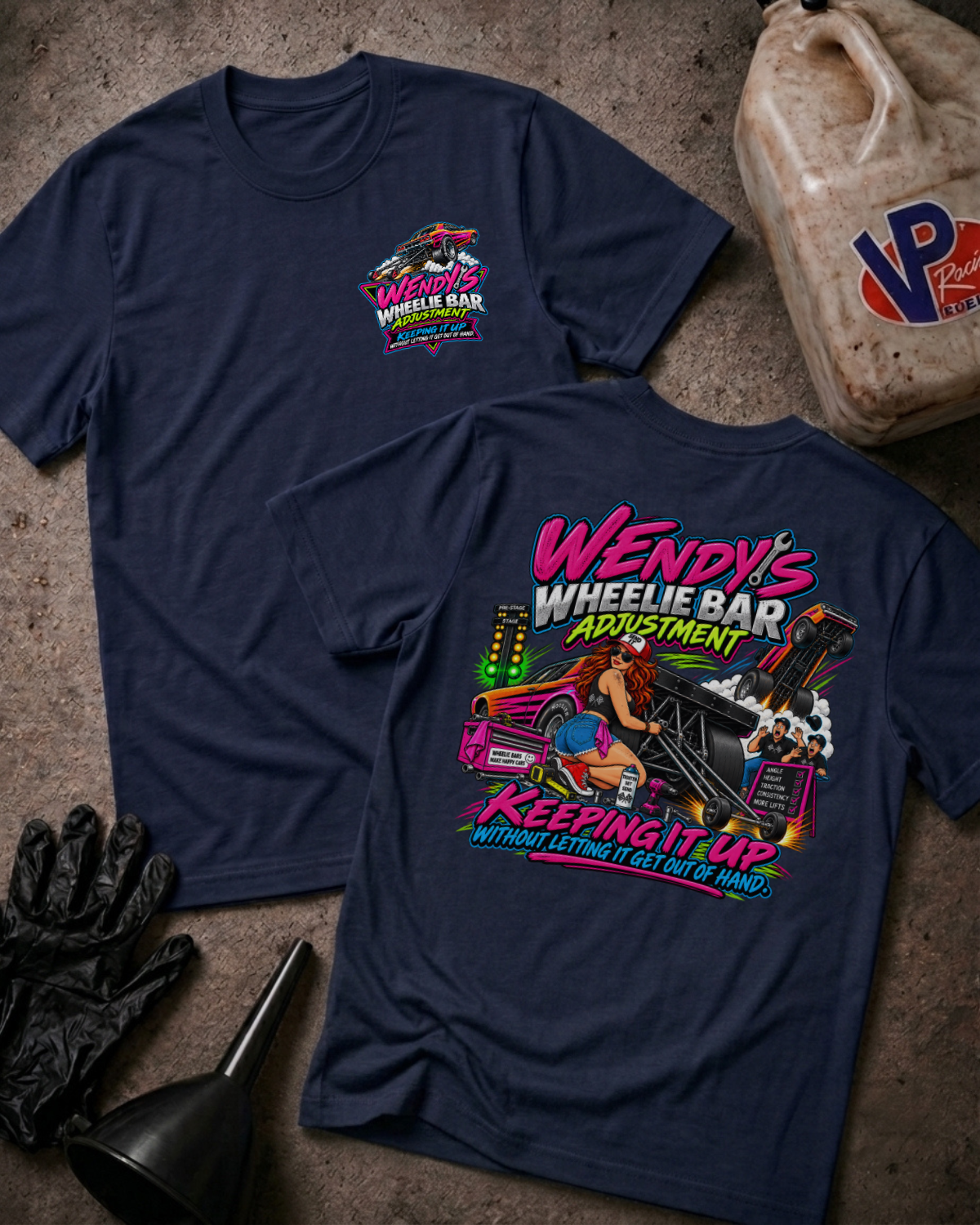 Two navy graphic t-shirts displayed on a textured concrete surface with garage-themed props including a plastic jug, black gloves, and a funnel. The front shirt shows a small pink and blue logo on the left chest, while the back shirt features a large colorful graphic of a woman in a pink car with the text 'Wendy's Wheelie Bar Adjustment Keeping It Up'.