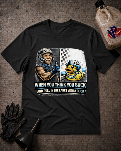 A black graphic t-shirt laid flat on a textured dark gray surface, styled with a race car driver, a duck in a helmet, and the text 'WHEN YOU THINK YOU SUCK AND PULL IN THE LANES WITH A DUCK'. A bottle, gloves, and funnel are placed around it as props.
