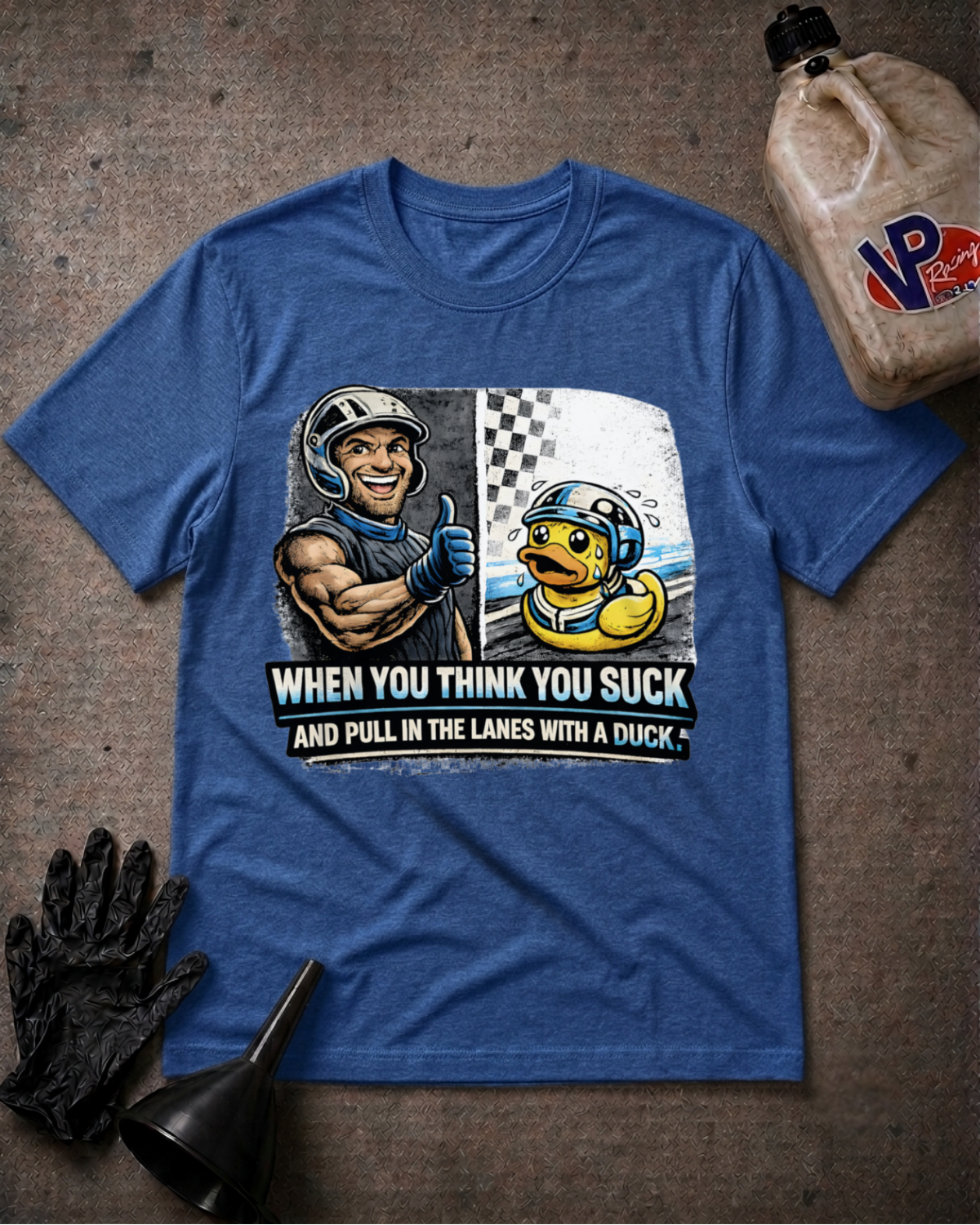 A heather blue graphic t-shirt laid flat on a textured gray surface, surrounded by racing-themed props including a black glove, funnel, and a jug of VP Racing fluid. The shirt features a graphic of a muscular racer giving a thumbs up next to a duck in a racing helmet with the text 'WHEN YOU THINK YOU SUCK AND PULL IN THE LANES WITH A DUCK'.