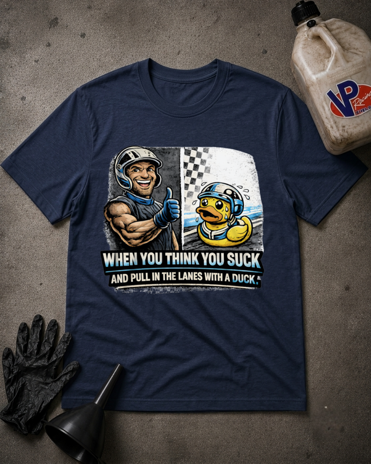 A navy blue graphic t-shirt laid flat on a textured gray surface, surrounded by racing-themed props including a bottle of VP racing oil, black gloves, and a funnel. The shirt features a graphic of a muscular racecar driver giving a thumbs up next to a yellow duck in a racing helmet, with the text 'WHEN YOU THINK YOU SUCK AND PULL IN THE LANES WITH A DUCK'.