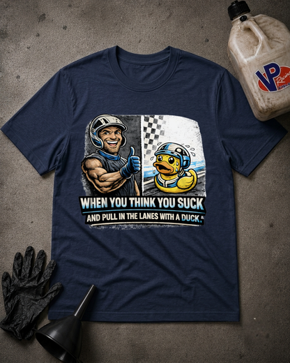 A navy blue graphic t-shirt laid flat on a textured gray surface, surrounded by racing-themed props including a bottle of VP racing oil, black gloves, and a funnel. The shirt features a graphic of a muscular racecar driver giving a thumbs up next to a yellow duck in a racing helmet, with the text 'WHEN YOU THINK YOU SUCK AND PULL IN THE LANES WITH A DUCK'.