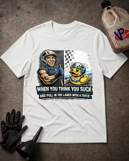 A white graphic t-shirt laid flat on a textured gray surface, surrounded by racing-themed props including a black glove, funnel, and a plastic jug. The shirt features a graphic of a muscular race car driver giving a thumbs up next to a yellow duck, with the text 'WHEN YOU THINK YOU SUCK AND PULL IN THE Lanes WITH A DUCK'.