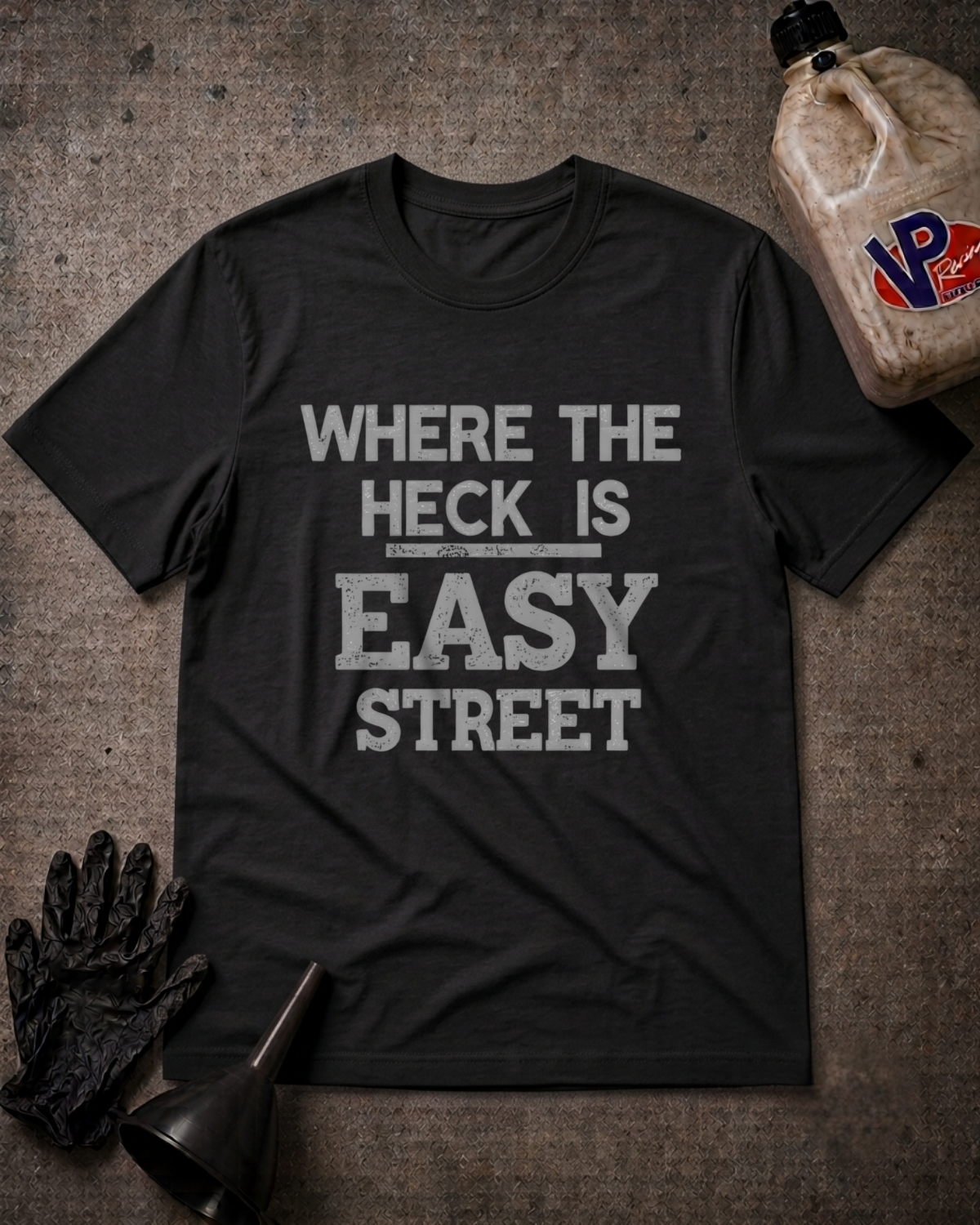 A black graphic t-shirt with the text 'WHERE THE HECK IS EASY STREET' printed in distressed white letters, laid flat on a textured gray background with automotive-themed props including a bottle, gloves, and a funnel.