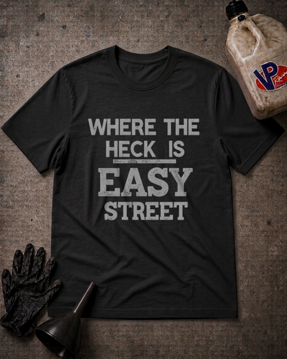 A black graphic t-shirt with the text 'WHERE THE HECK IS EASY STREET' printed in distressed white letters, laid flat on a textured gray background with automotive-themed props including a bottle, gloves, and a funnel.