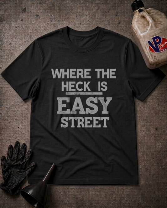 A black graphic t-shirt with the text 'WHERE THE HECK IS EASY STREET' printed in distressed white letters, laid flat on a textured gray background with automotive-themed props including a bottle, gloves, and a funnel.
