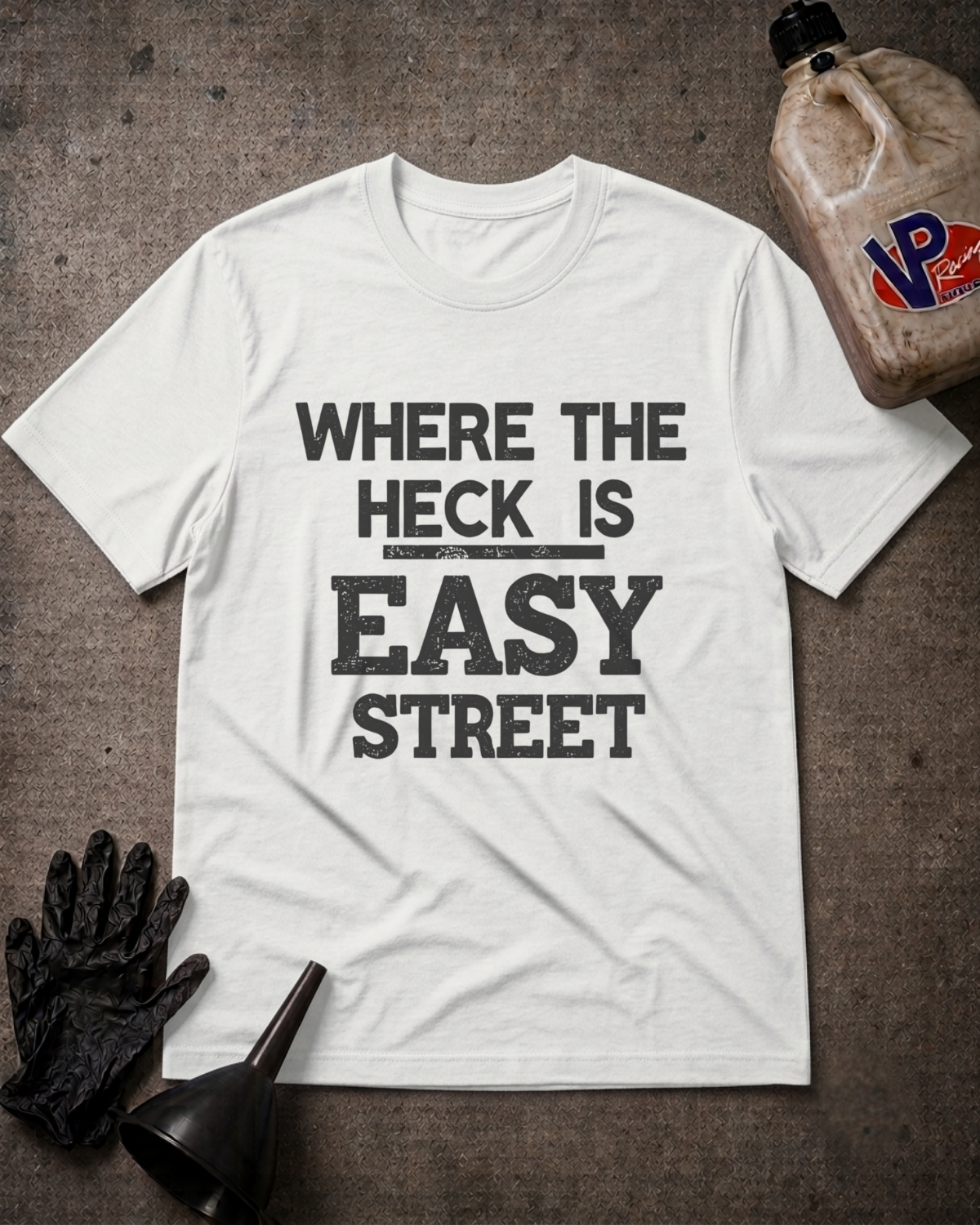 A white graphic t-shirt with the text 'WHERE THE HECK IS EASY STREET' printed in distressed black lettering. The shirt is displayed in a flat-lay arrangement on a dark textured background, accompanied by a black work glove, a black funnel, and a dirty plastic jug of oil with a 'VP' logo.