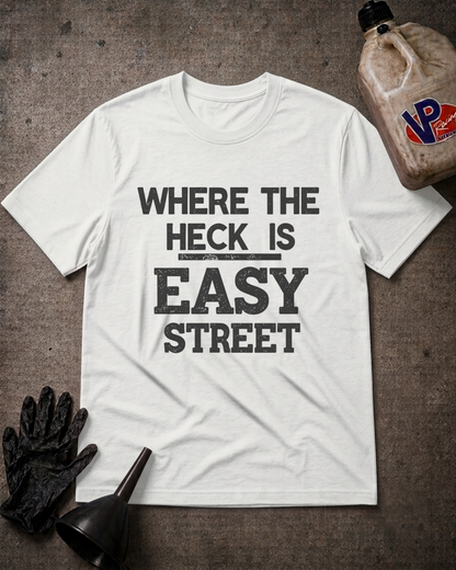 A white graphic t-shirt with the text 'WHERE THE HECK IS EASY STREET' printed in distressed black lettering. The shirt is displayed in a flat-lay arrangement on a dark textured background, accompanied by a black work glove, a black funnel, and a dirty plastic jug of oil with a 'VP' logo.