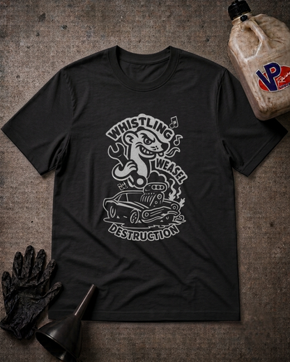 A black graphic t-shirt featuring a white cartoon illustration of a weasel driving a wrecked car with the text 'WHISTLING WEASEL DESTRUCTION'. The t-shirt is displayed in a flat-lay arrangement on a textured gray background, surrounded by automotive-themed props including a black oil filter wrench, a black glove, and a plastic jug of VP Fluid.