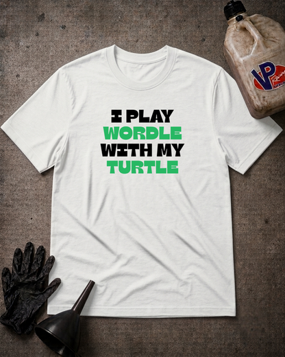 A white short-sleeve graphic T-shirt laid flat on a dark textured surface, with black and bright green text reading 'I PLAY WORDLE WITH MY TURTLE.' Black gloves and a black funnel sit at the lower left, and a plastic jug appears at the upper right as styled props.