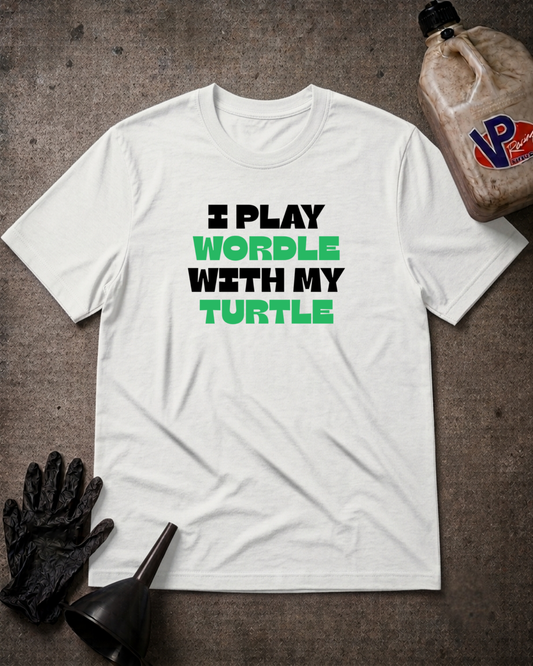 A white short-sleeve graphic T-shirt laid flat on a dark textured surface, with black and bright green text reading 'I PLAY WORDLE WITH MY TURTLE.' Black gloves and a black funnel sit at the lower left, and a plastic jug appears at the upper right as styled props.