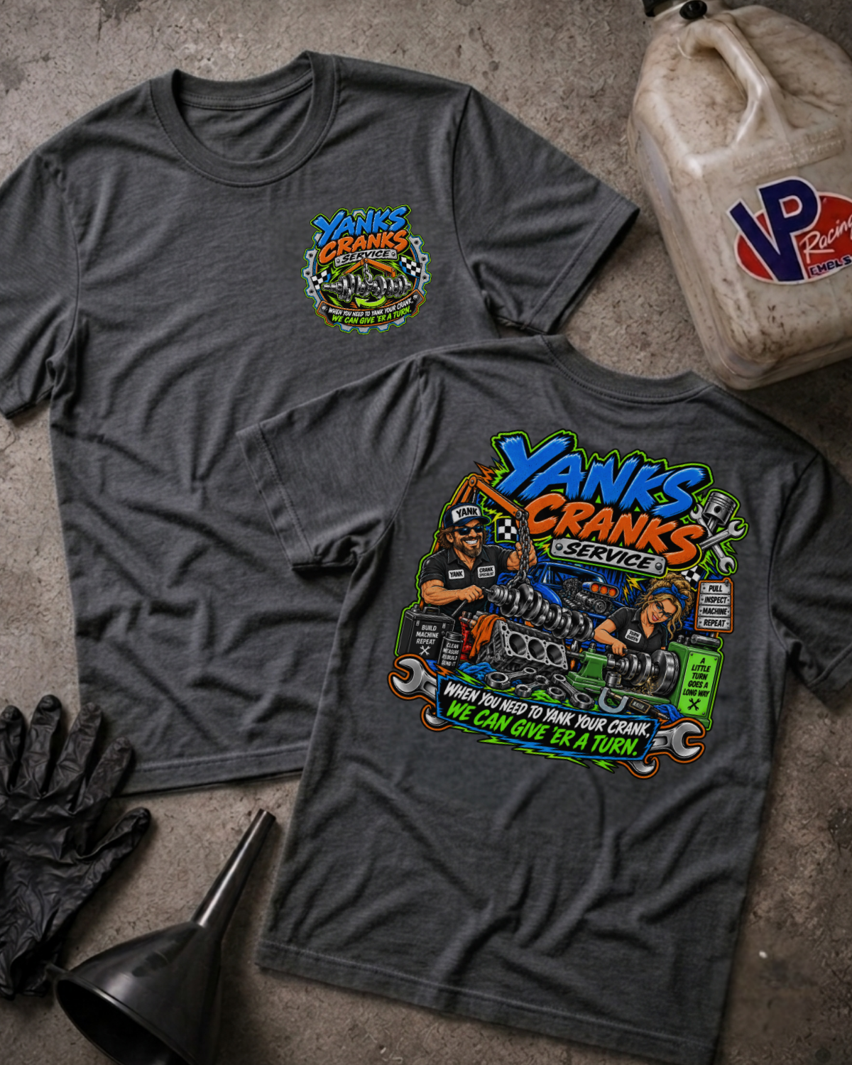 Two dark heather gray graphic T-shirts laid flat on a concrete surface, with a black funnel, black gloves, and a plastic oil container nearby. The front shirt shows a large colorful 'Yanks Cranks Service' graphic, and the back shirt shows a smaller matching chest graphic.